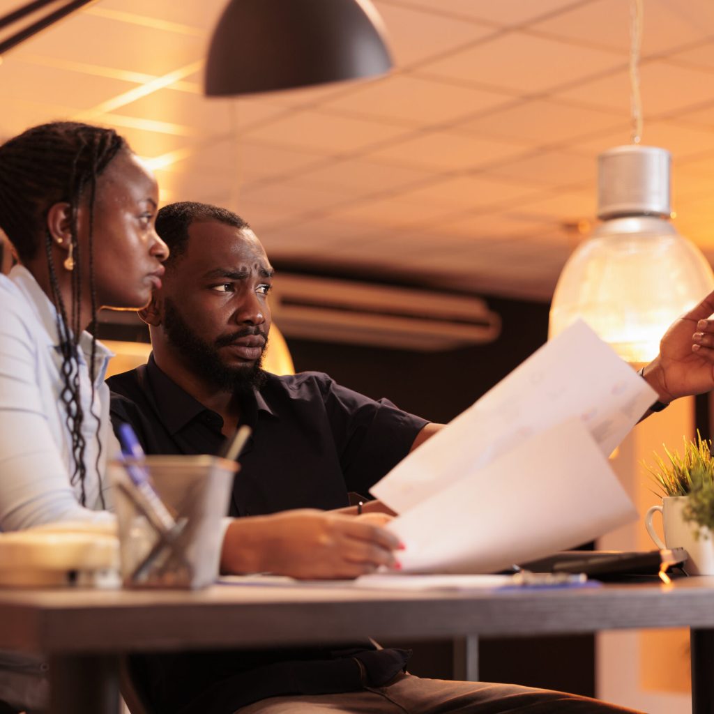 African american man and woman doing teamwork collaboration African american man and woman doing teamwork collaboration to do presentation report, looking at data on computer and papers. Working together with paperwork in office during sunset.